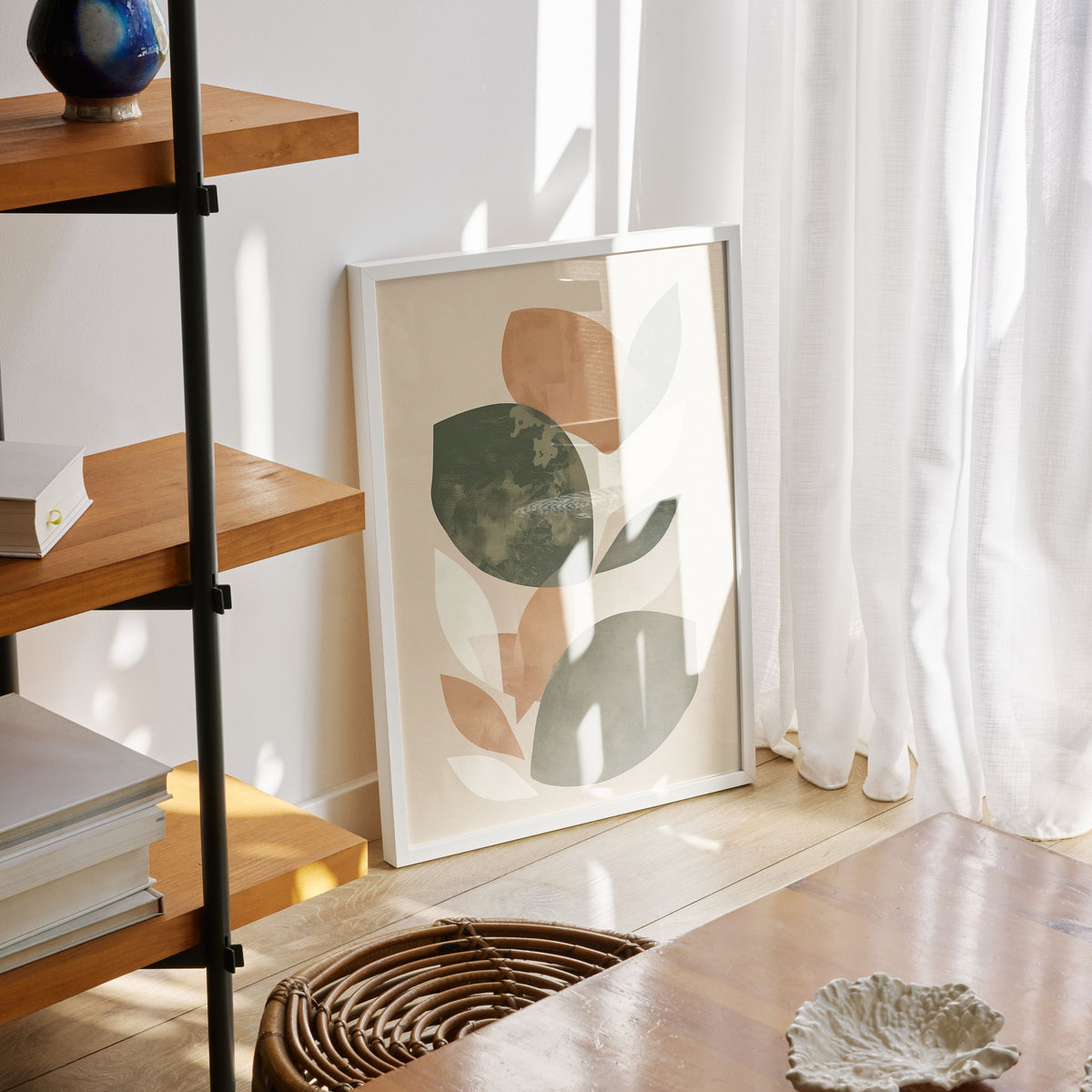Minimalist abstract botanical print with soft green, beige, and muted peach shapes framed in white, leaning against a sunlit wall near sheer curtains and wooden furniture.