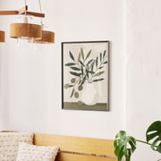 Framed botanical wall art featuring abstract olive branch design in muted green tones, hanging above a wooden bench with textured cushions in a bright, minimal room.