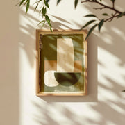 Abstract botanical wall art print featuring earthy olive green and beige shapes, framed in natural wood, with soft sunlight and leafy shadows enhancing the calming mood.