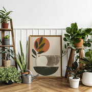 Framed abstract botanical wall art print with earthy tones, surrounded by various green houseplants and a metal watering can on a wooden floor against a white paneled wall.