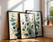 Three framed botanical art prints with eucalyptus leaves and small yellow flowers, leaning against a wall on a wooden floor next to a window with sunlight and a potted plant.