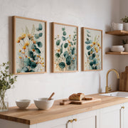 Three framed botanical wall art prints with teal eucalyptus leaves and yellow flowers hang above a kitchen counter with bowls, bread, and a gold faucet.