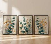 Three framed botanical art prints with eucalyptus leaves and yellow-orange wildflowers, leaning against a beige wall on a light wood floor.