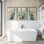 Three framed botanical wall art prints with green eucalyptus leaves and yellow flowers hang above a white freestanding bathtub in a bright bathroom.