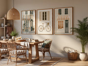 Three framed wall art prints depicting classic street scenes and a bicycle, styled in a warm, modern dining room with a wooden table, woven chairs, and a potted plant.