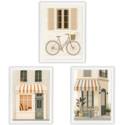 Three framed art prints featuring Parisian-style storefront scenes, including a bicycle on a cream building, a shop with a striped awning, and a cafe exterior with chairs and plants.